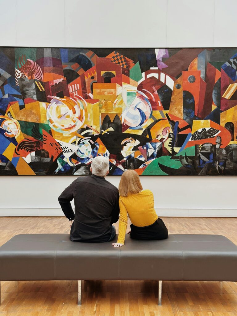 A couple sits in a modern gallery, captivated by an abstract painting full of vibrant colors.